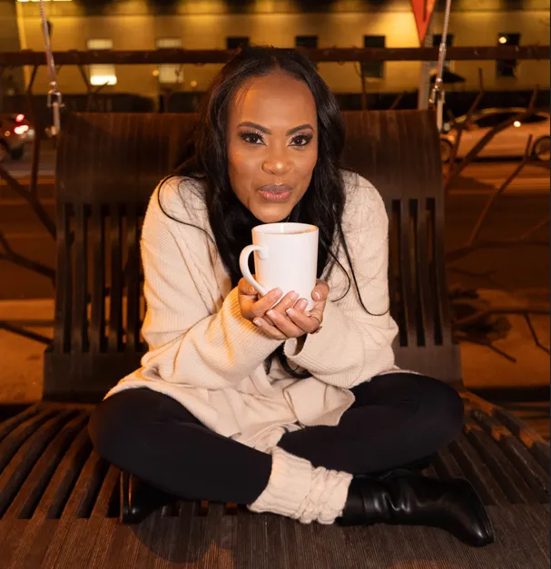 Rosa Allison sitting cross-legged on a wooden bench at night in an oversized cream cardigan, holding a white mug in both hands.